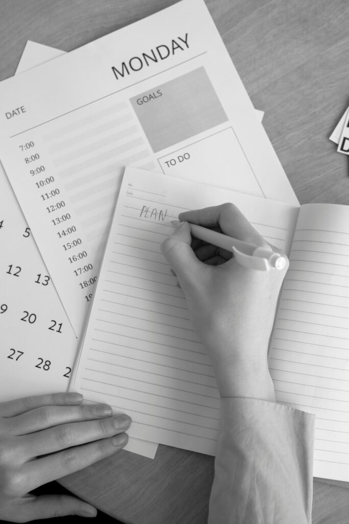 A monochrome image of a person planning their week with a notebook, calendar, and goals list.